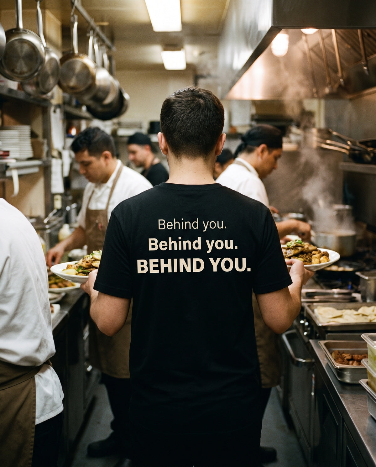 Behind You Tee worn in a busy restaurant kitchen, back print in cream on black