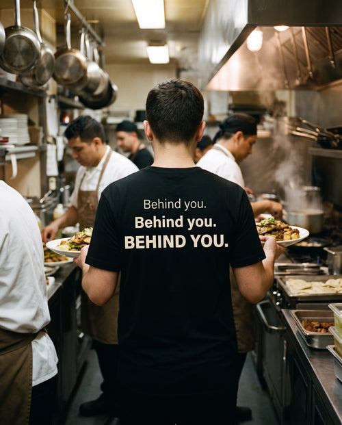 Behind You Tee worn in a busy restaurant kitchen, back print in cream on black