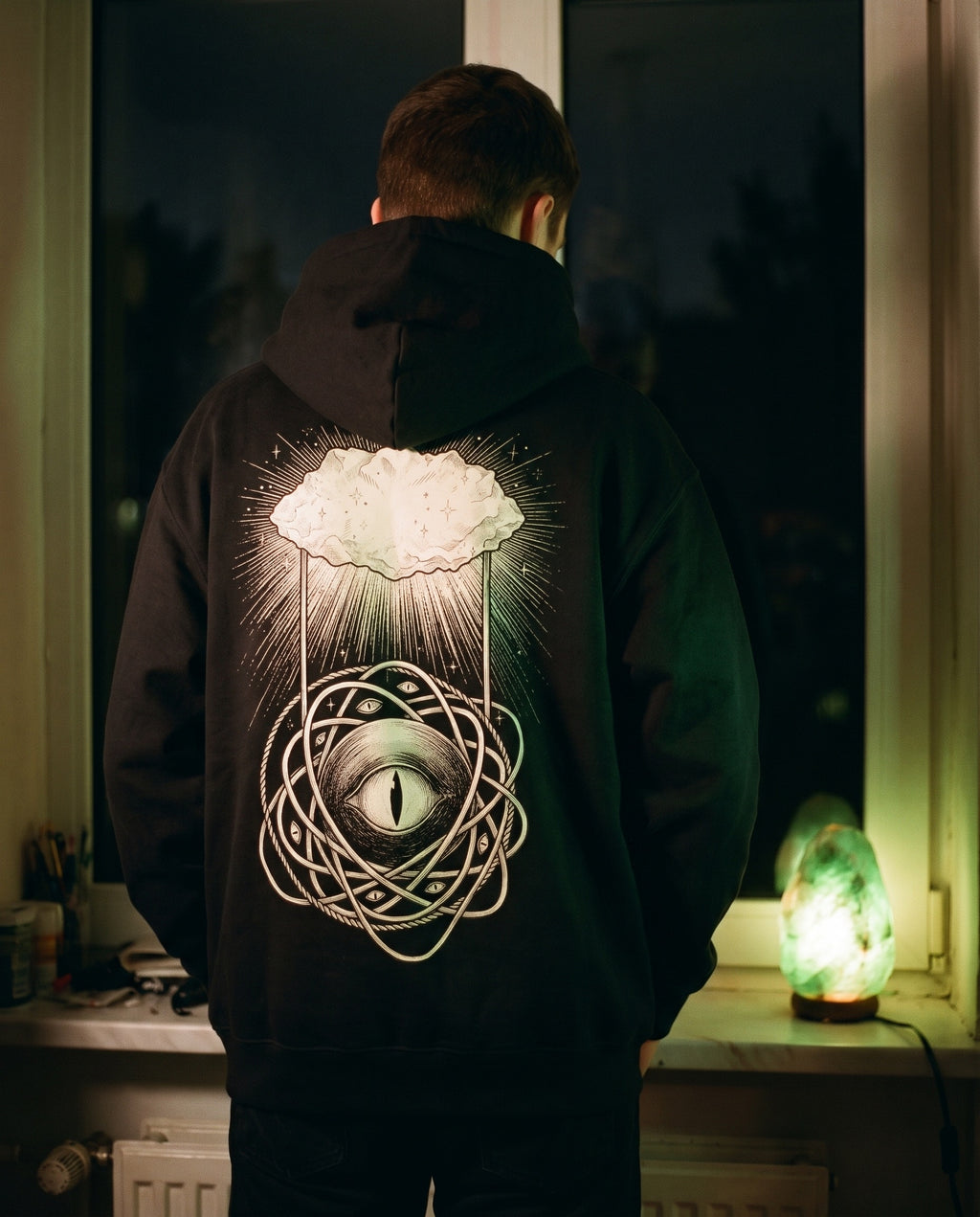 Jaded Hoodie back print, worn by a window lit by a green crystal